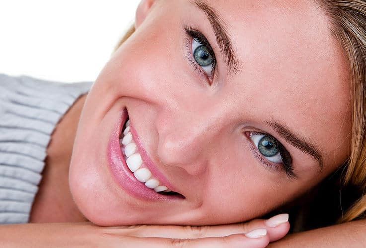 Woman with blonde hair smiling at camera, resting head on hand against white background.
