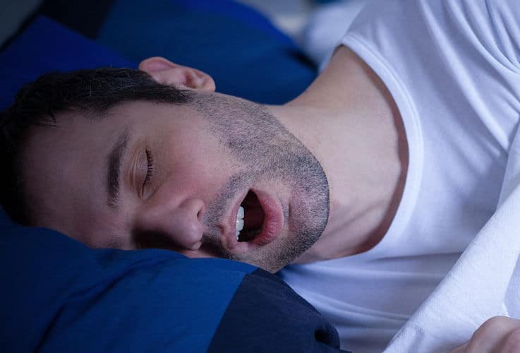 Man sleeping peacefully on blue pillow wearing white shirt, head tilted to side.