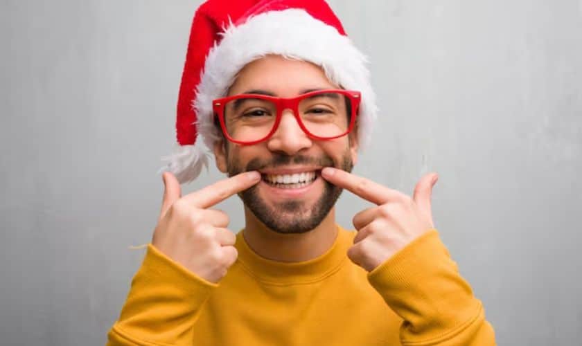 Smiling man in Santa hat and glasses pointing at cheeks against gray background.