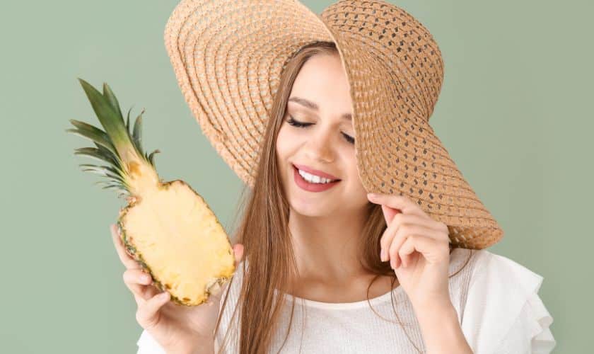 Woman holding pineapple wearing straw hat against green background, smiling.