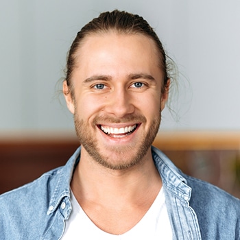 Smiling man wearing denim shirt over white t-shirt, sitting indoors against neutral background.
