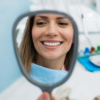 Woman smiling at mirror in dental office, wearing bib, showing healthy teeth.