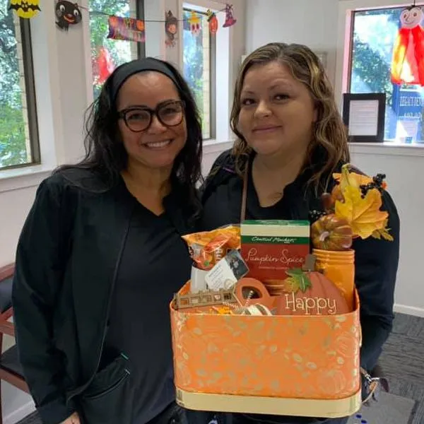 Two women smiling at camera next to decorated fall-themed cake in classroom setting.