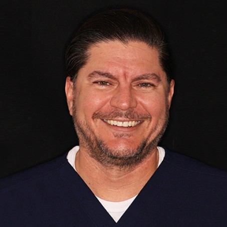 Man with dark hair and beard smiling at camera against black background, wearing dark blue shirt.