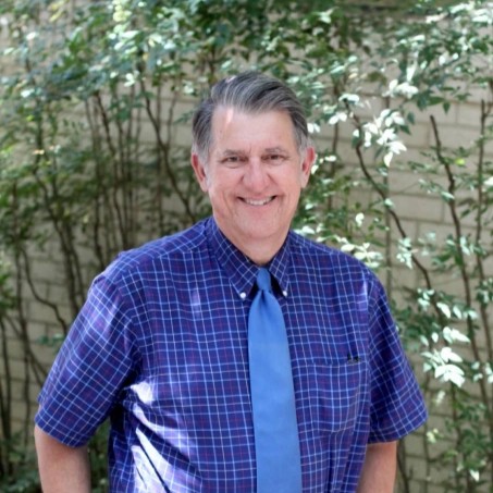 Middle-aged man in blue plaid shirt and tie smiling outdoors against foliage background.